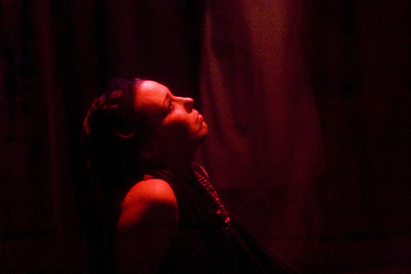 uma mulher, vista de perfil, olha para cima, banhada por uma luz vermelha, em um ambiente escuro